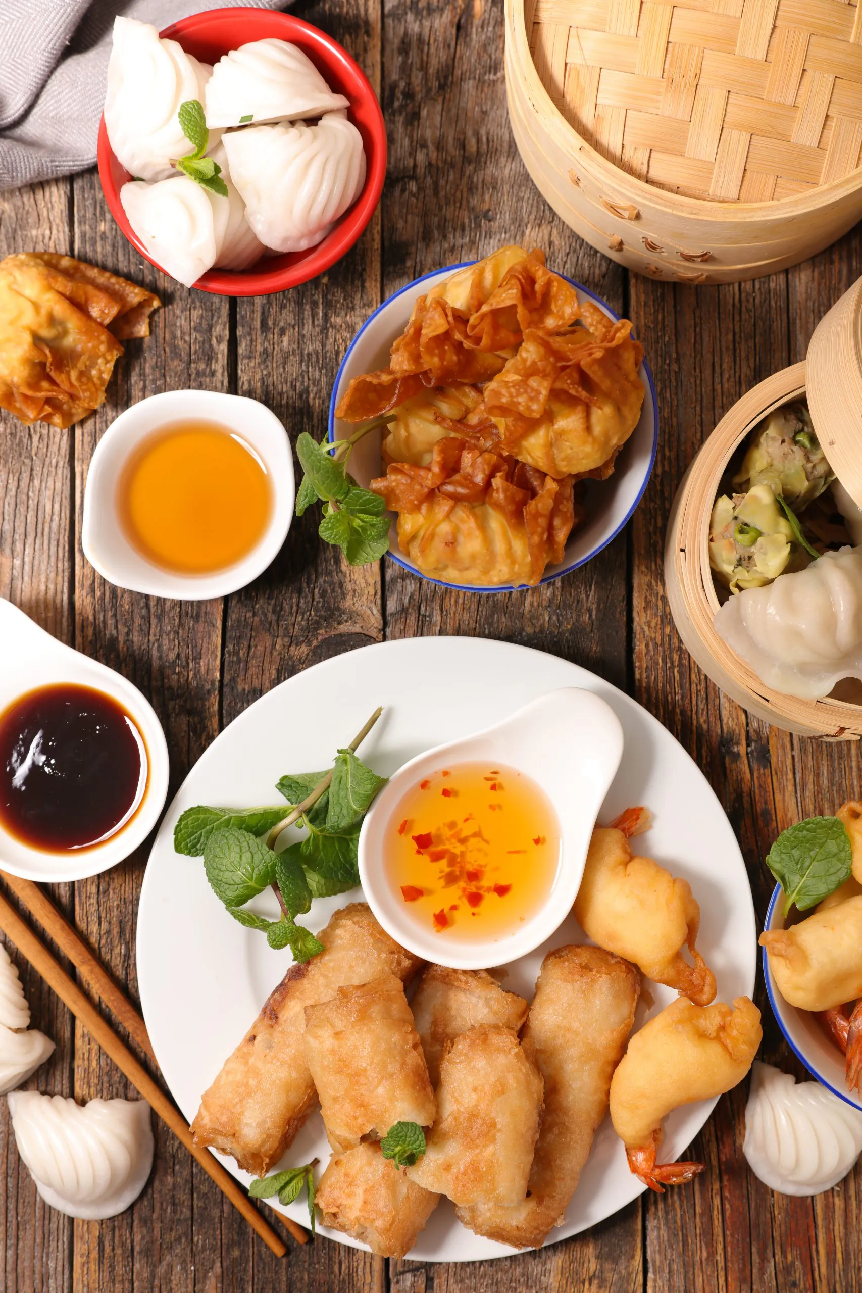 A rustic wooden table laden with assorted Chinese dim sum: steamed dumplings, fried spring rolls, crispy wontons, shrimp toast, and dipping sauces—garnished with fresh mint.